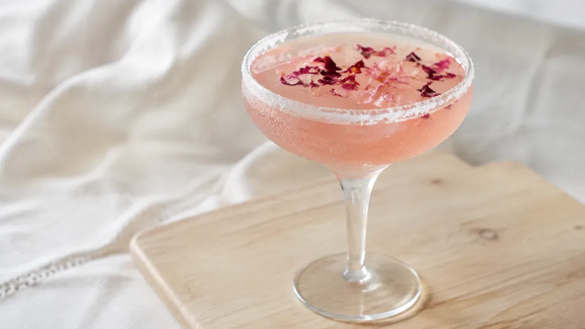 Copa de margarita rosada decorada con pétalos de rosa, sobre una mesa de madera con luz natural
