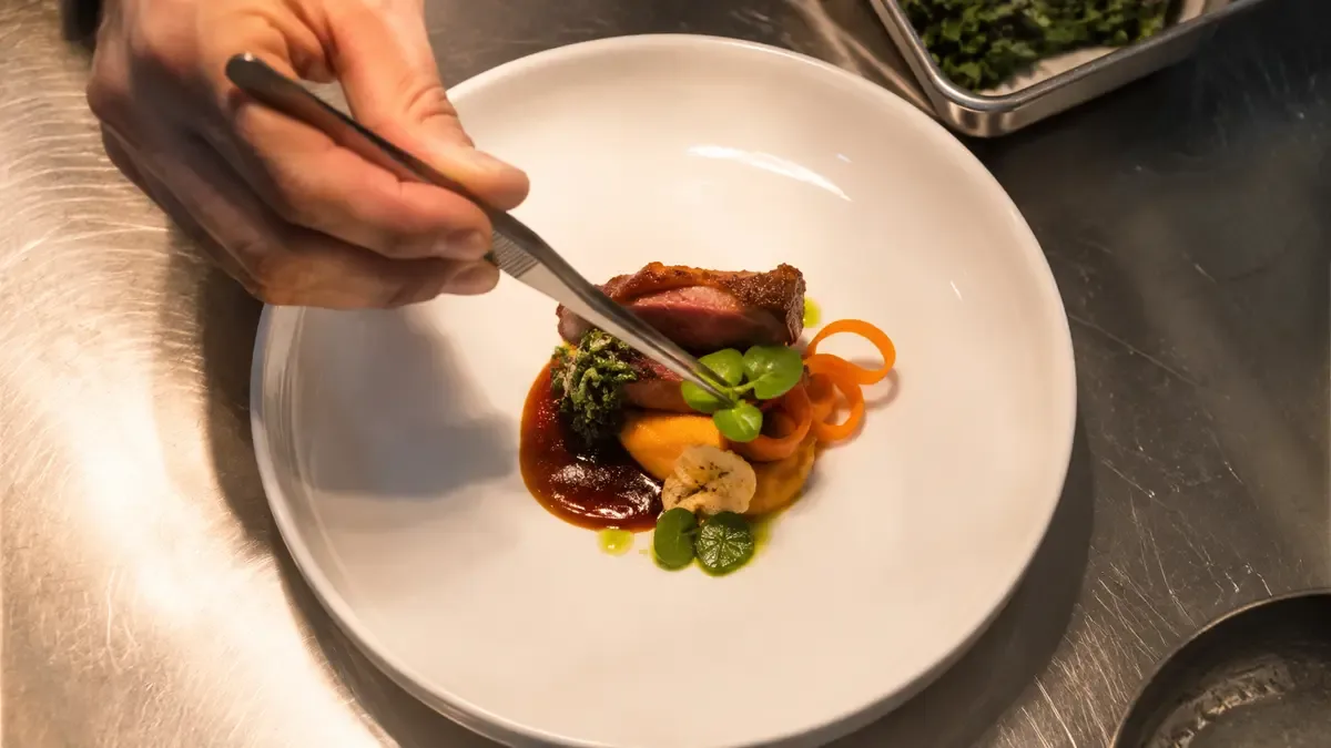 Mano con pinza colocando un detalle sobre un plato a medio emplatar