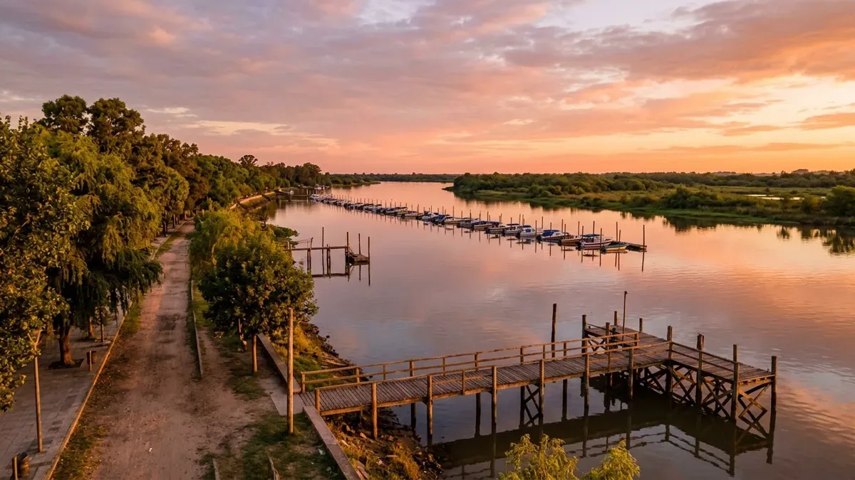 Costanera junto al río con árboles frondosos y un muelle de madera al atardecer, sin personas
