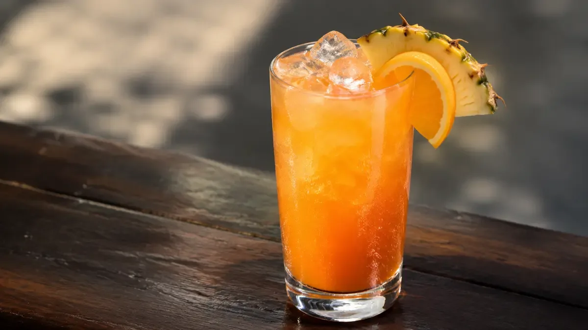 Cóctel tropical color naranja con rodajas de ananá y naranja sobre una mesa de madera